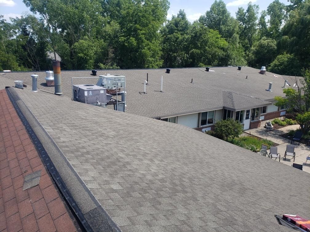 Flat rooftop with HVAC units and vents beside a red shingle roof, surrounded by trees