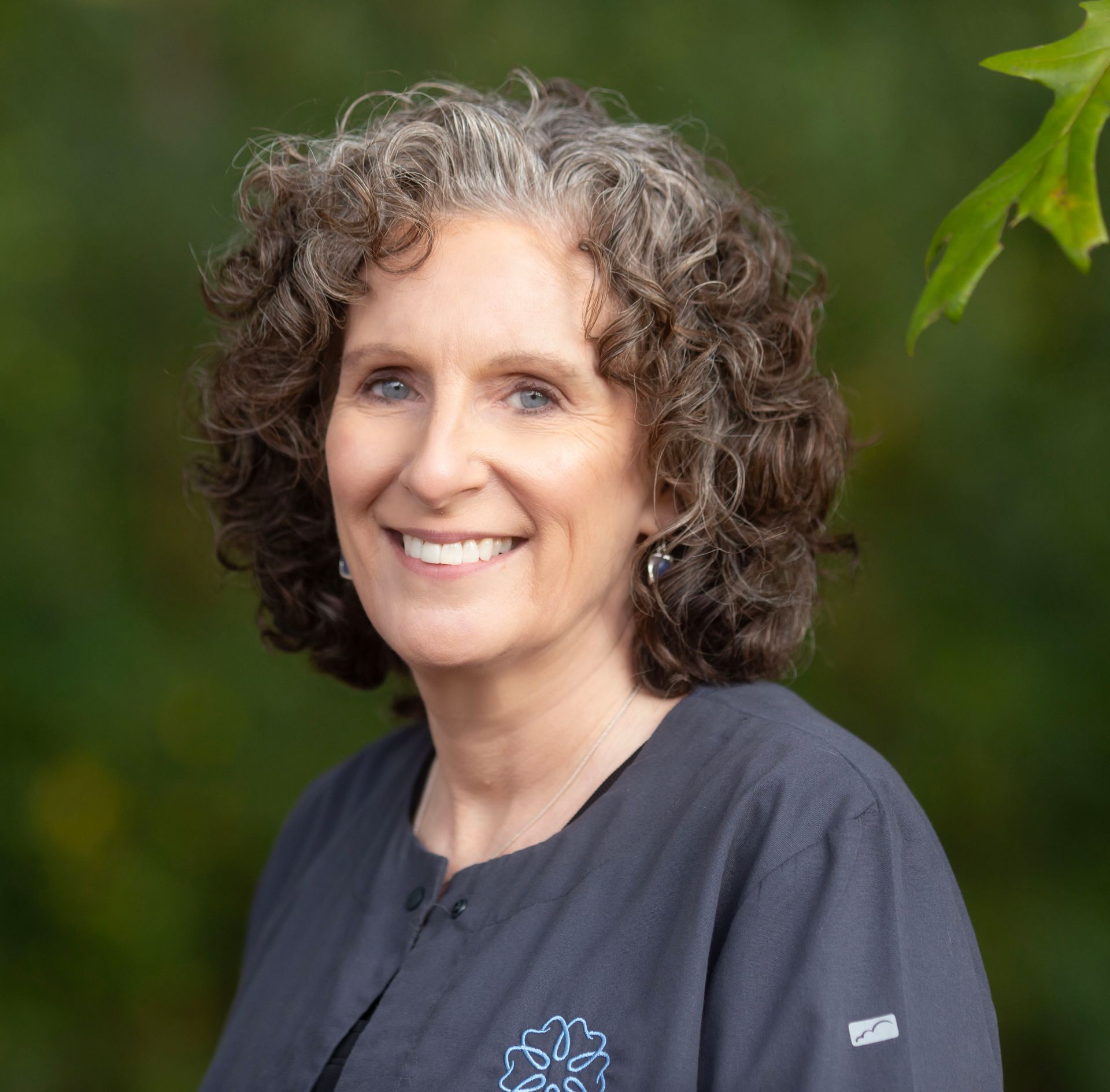 Smiling woman with curly gray hair in a dark shirt against a blurred green background