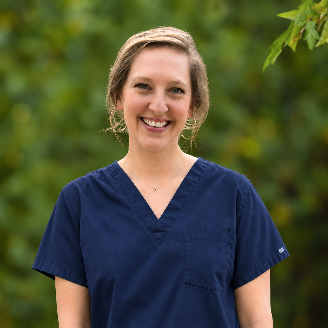 Smiling person in dark gray scrubs outdoors against a blurred green background