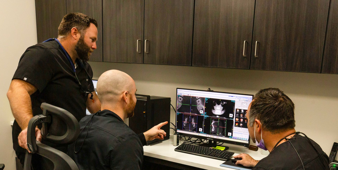 Three men in an office review content on a desktop computer with a lit screen.