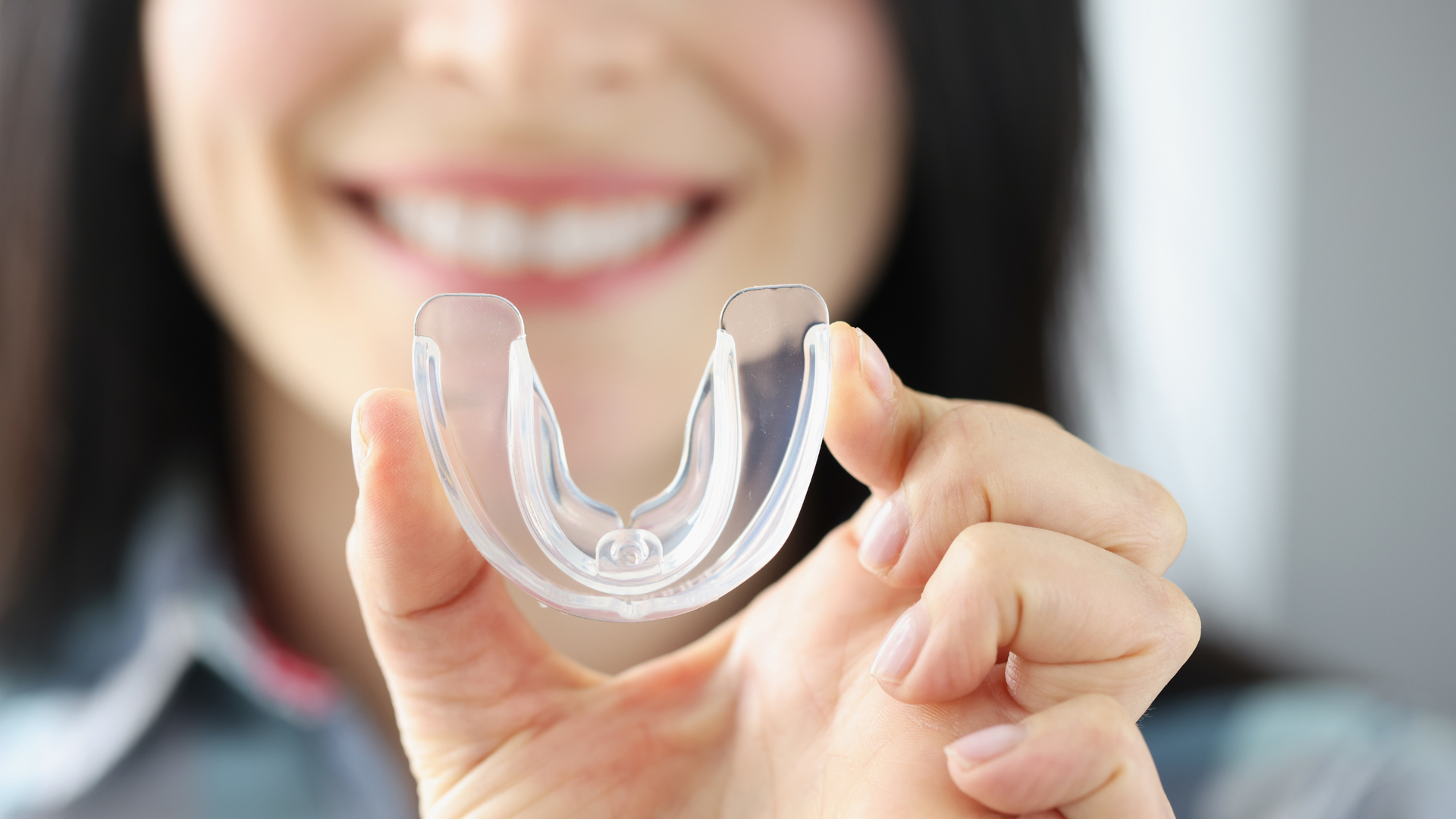 Woman smiling, holding clear dental retainer close to the camera.