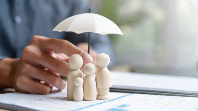 Hands arranging wooden figures under a white umbrella on a desk, symbolizing family insurance protection.