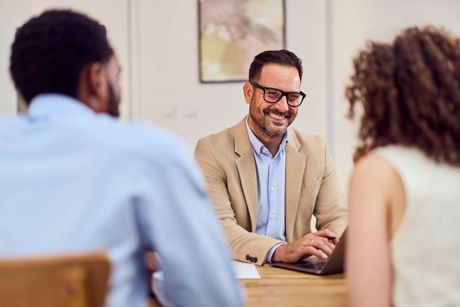 Two people meet with a smiling man at a desk in a bright office