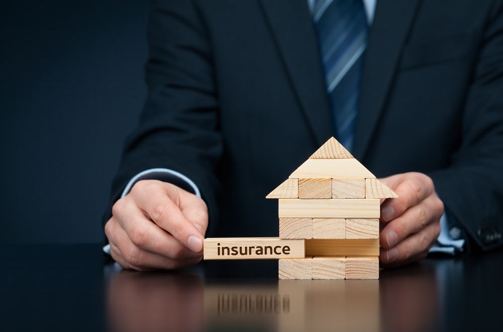 Hands in suit beside wooden house model and “insurance” block on a dark table