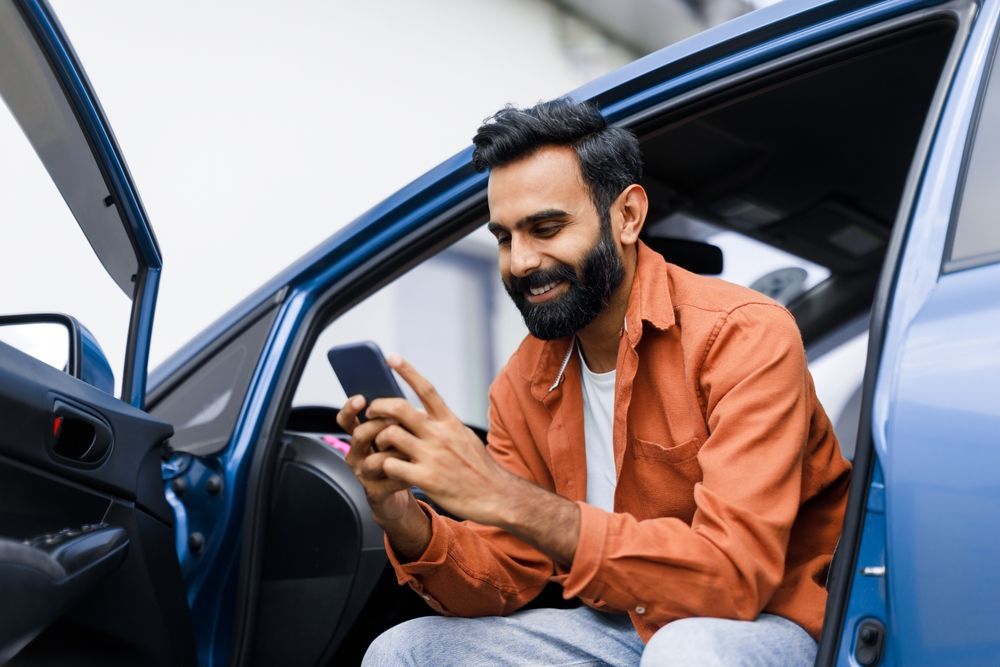 Man smiling while using a smartphone inside a blue car with the door open
