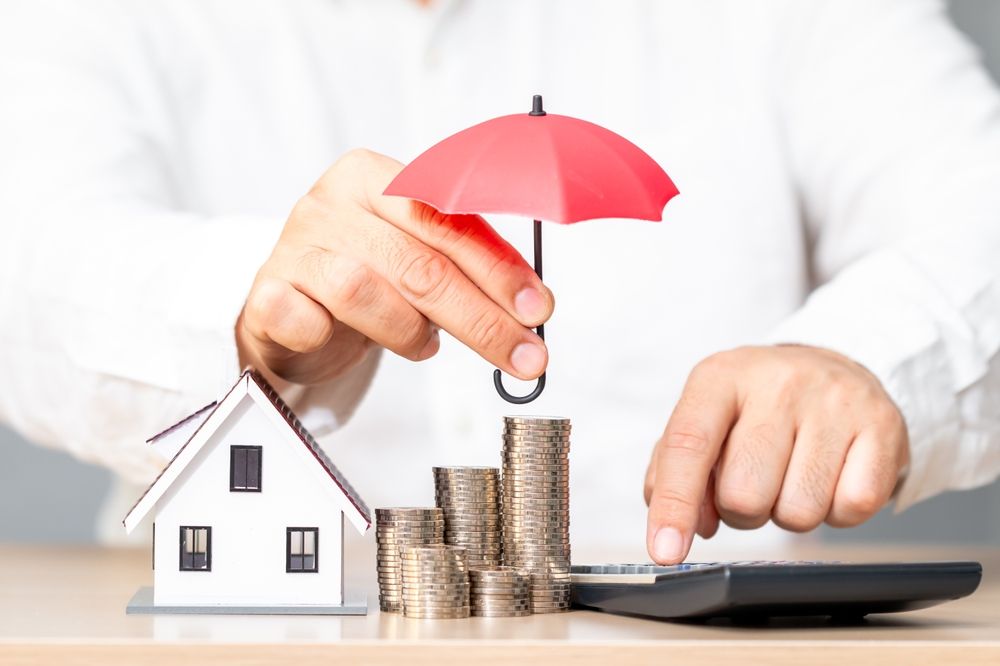 Hands placing red umbrella over house model, coins, and calculator on a desk