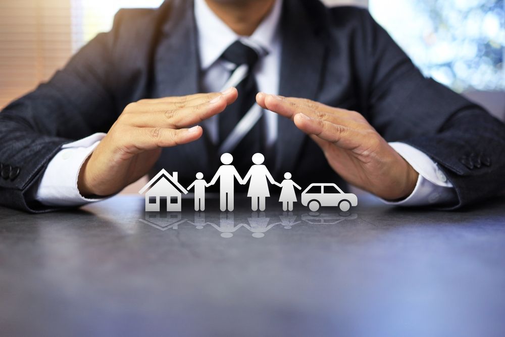 Hands protecting a family, house, and car icons on a table, symbolizing insurance protection