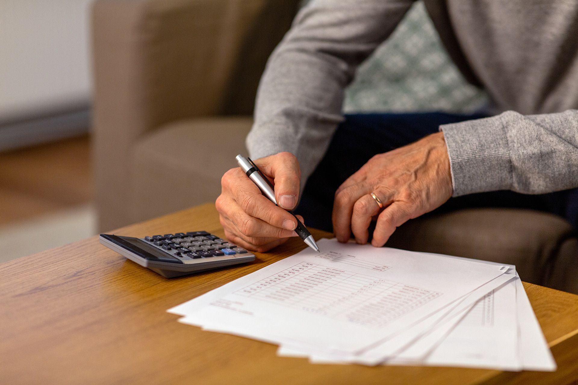 Person writing on papers beside a calculator on a wooden table with a laptop in the background