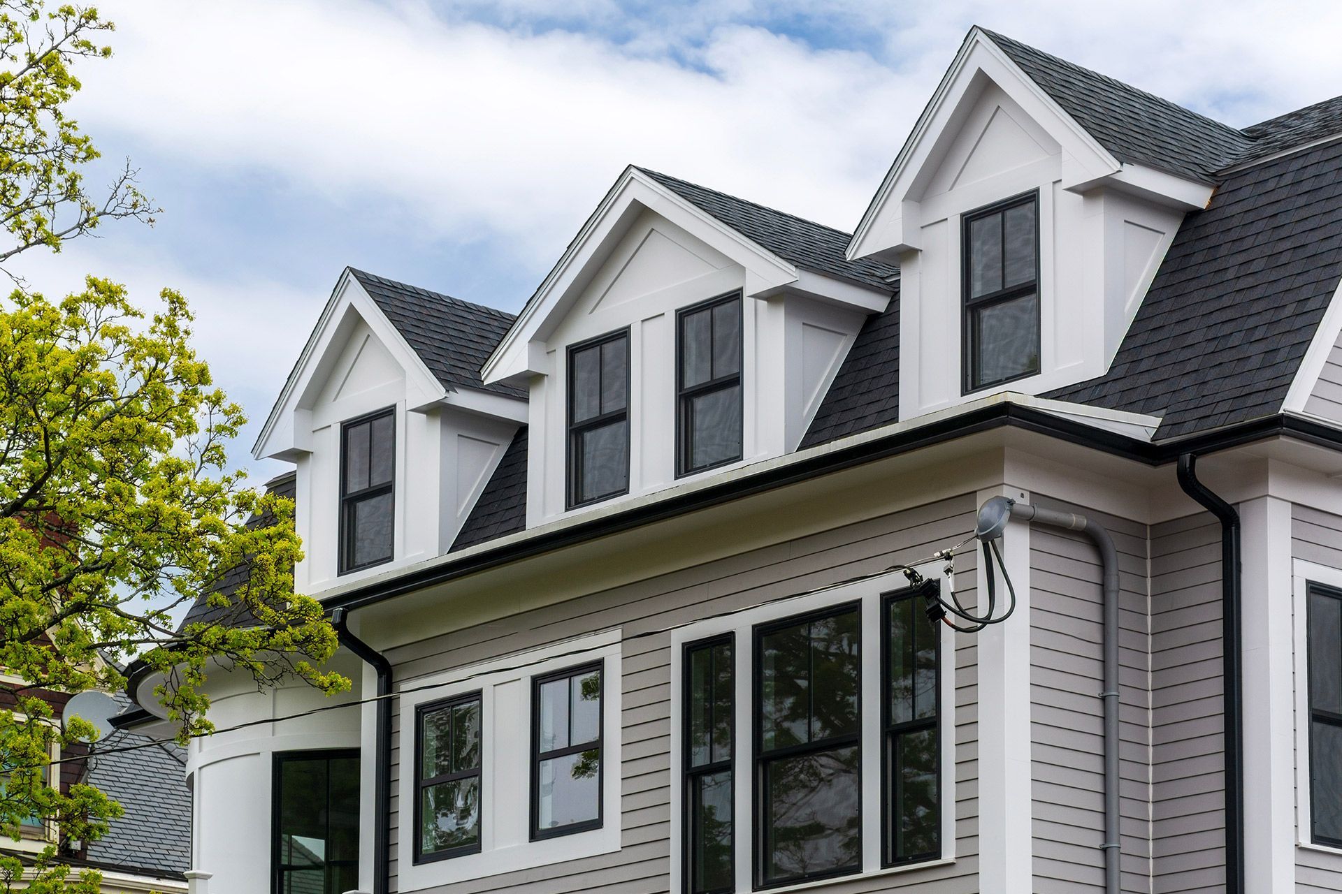 White house with black shutters and dormer windows, framed by green trees on a sunny day