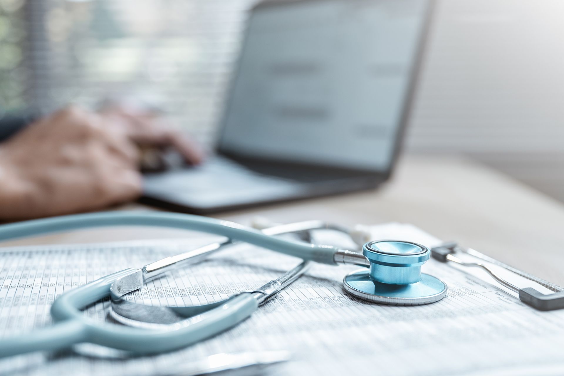 Blue stethoscope on a desk with a blurred person using a laptop in the background