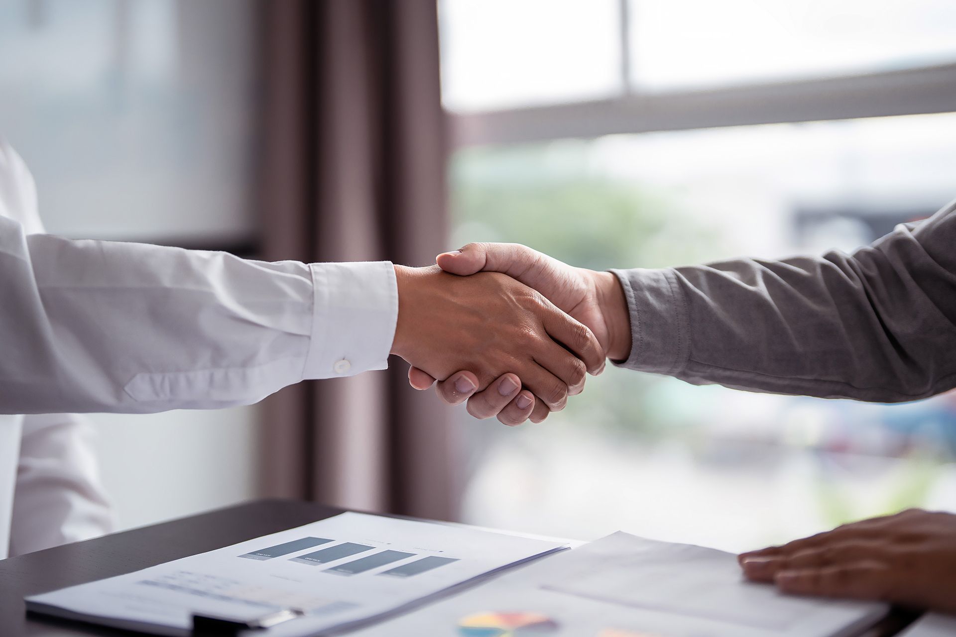 Two people shaking hands across a desk with documents in a bright office