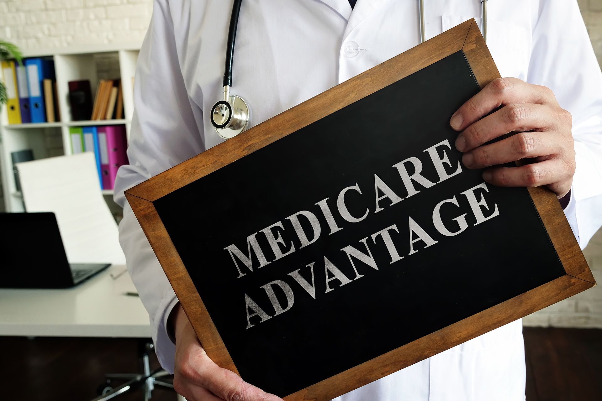 Doctor holding a chalkboard sign reading “MEDICARE ADVANTAGE” in a clinical office