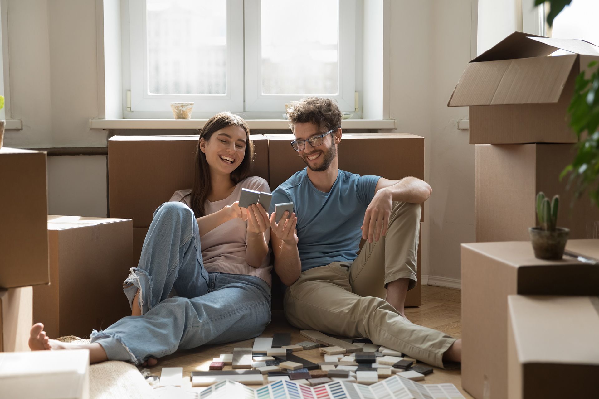 Couple sitting on the floor among moving boxes, smiling while looking at a smartphone in a sunlit room.