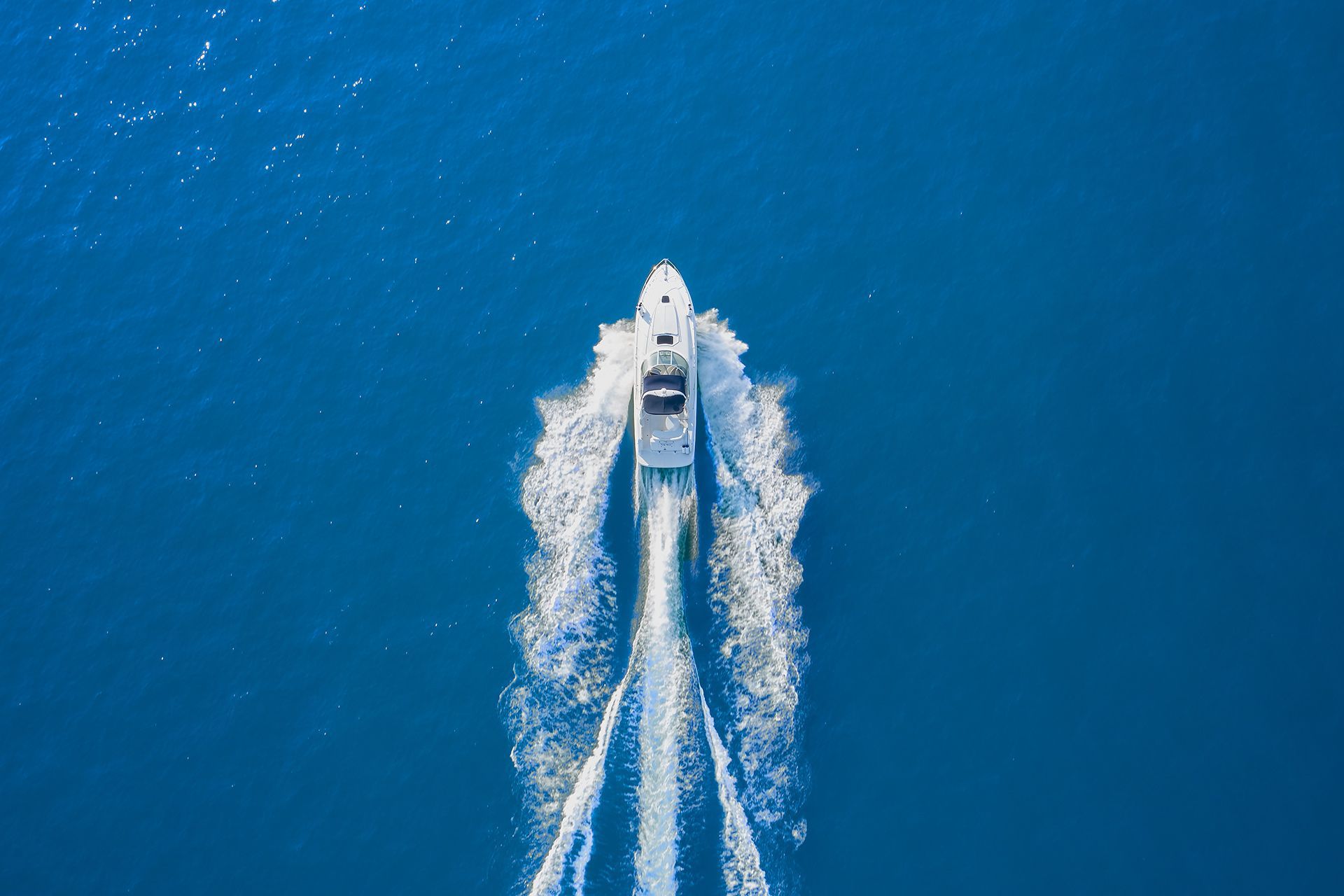 White speedboat cutting through deep blue water, leaving two foamy wakes from an aerial view