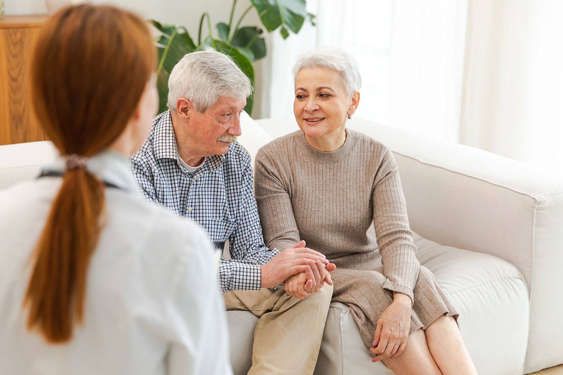 Three people talking on a white couch in a bright living room, one holding an older person’s hand.