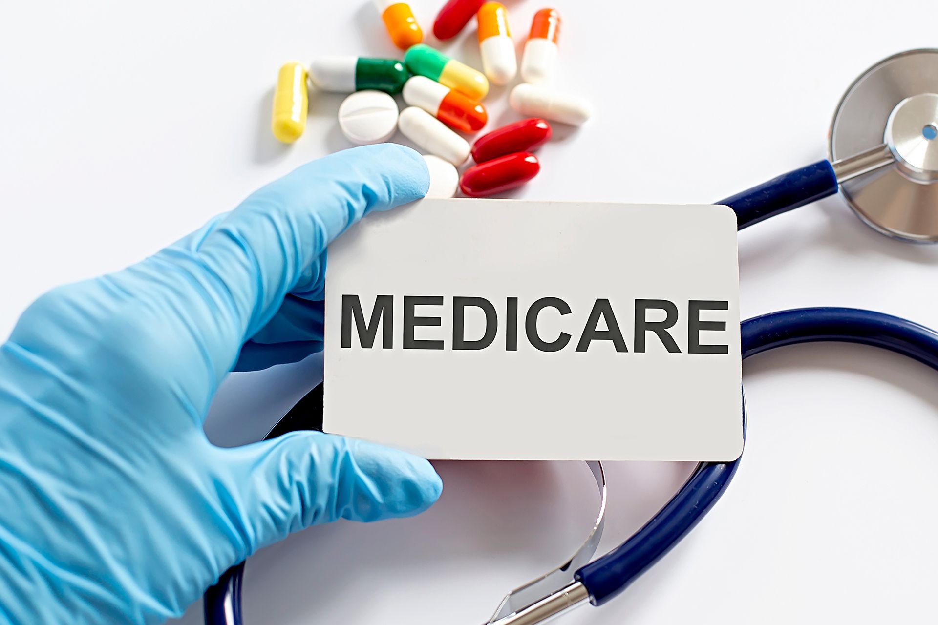 Gloved hand holding a Medicare card beside colorful pills and a stethoscope
