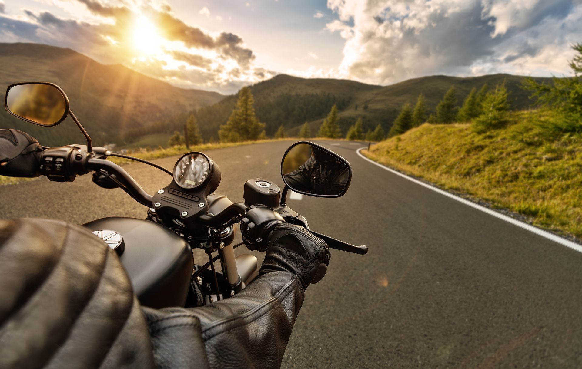 Motorcyclist riding a winding mountain road at sunset, with gloved hands on the handlebars.
