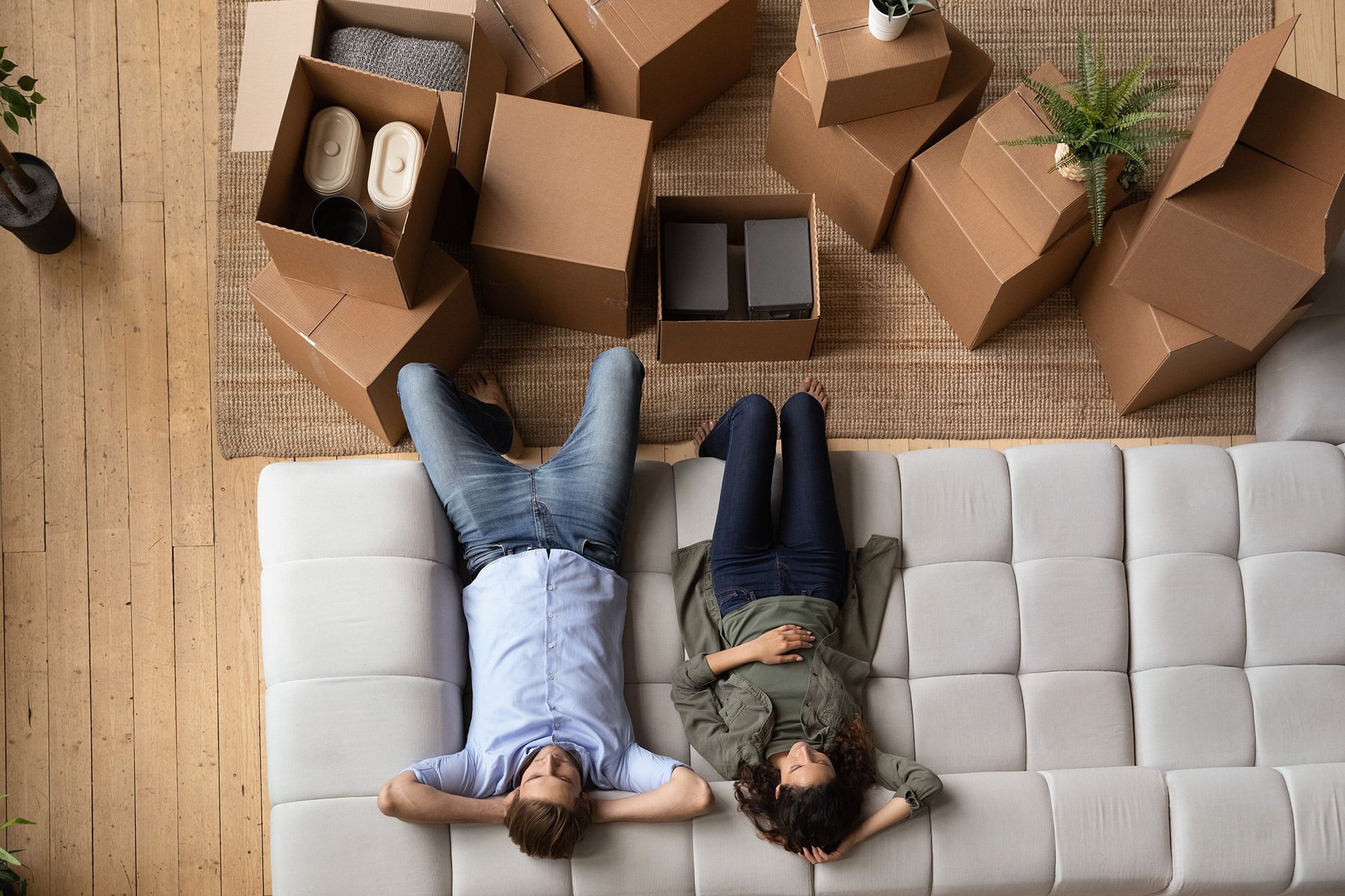 Two people lie on a white couch amid moving boxes and unpacked belongings in a room with wood floors.