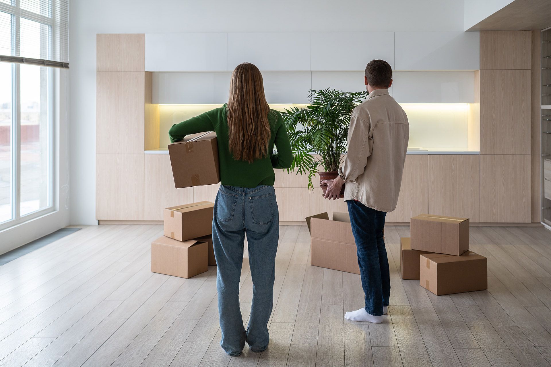 Two people carrying boxes in a bright, empty room with built-in shelving and a floor lamp.