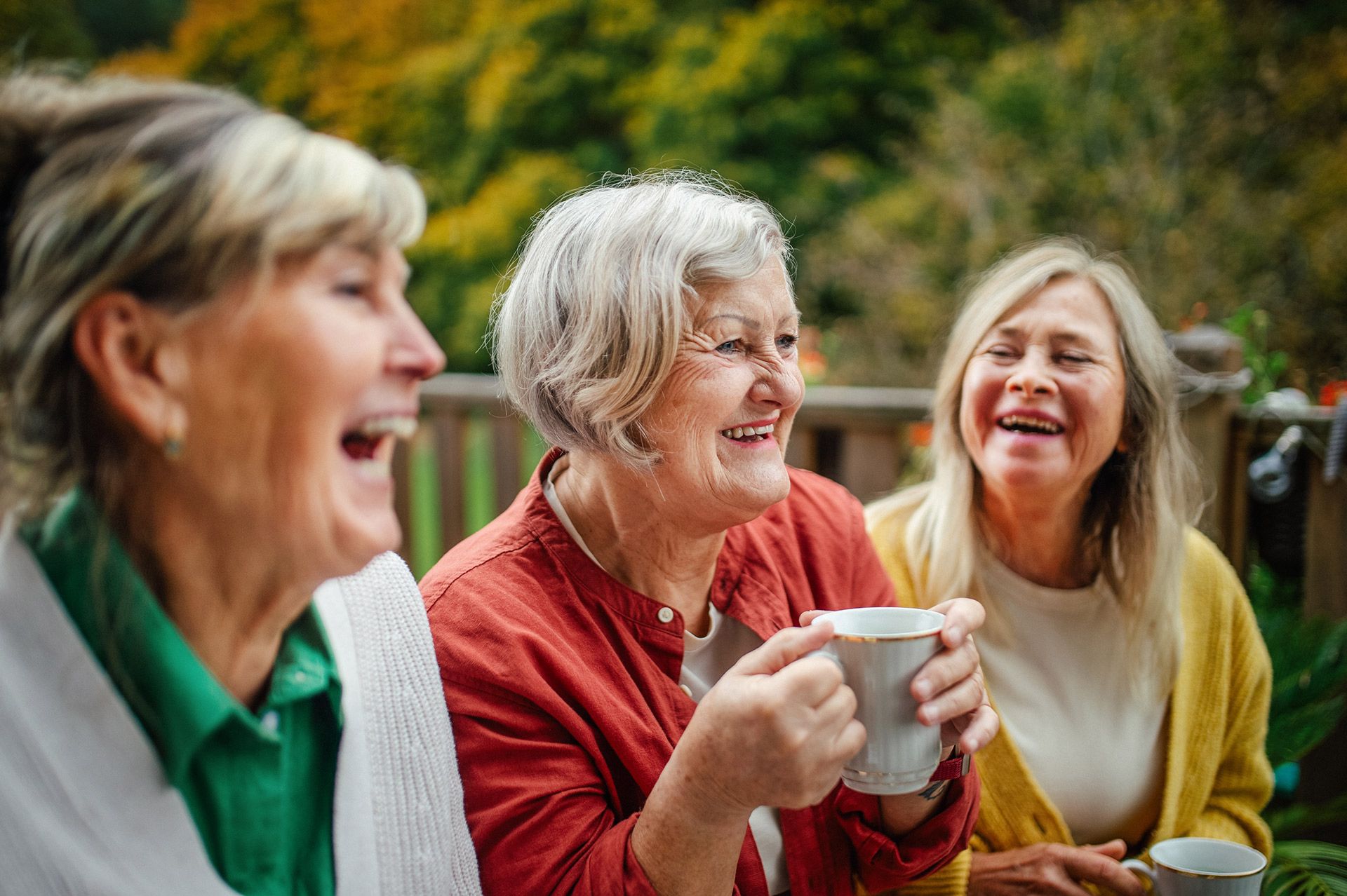 Three people laughing and holding cups outdoors in a garden setting