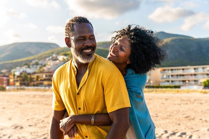 Couple smiling on a beach, with mountains and buildings in the background.
