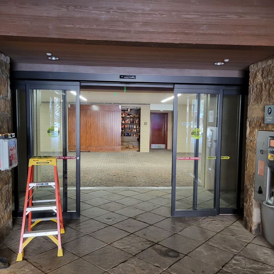 Automatic sliding glass doors open, revealing a hallway. A ladder stands in front of the doors.