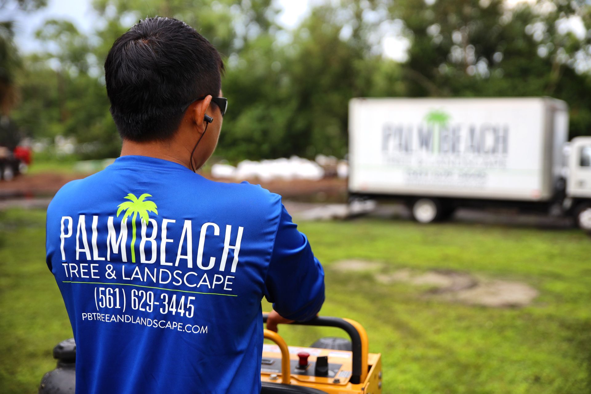 Man in blue Palm Beach Tree & Landscape shirt mowing grass beside a white truck outdoors