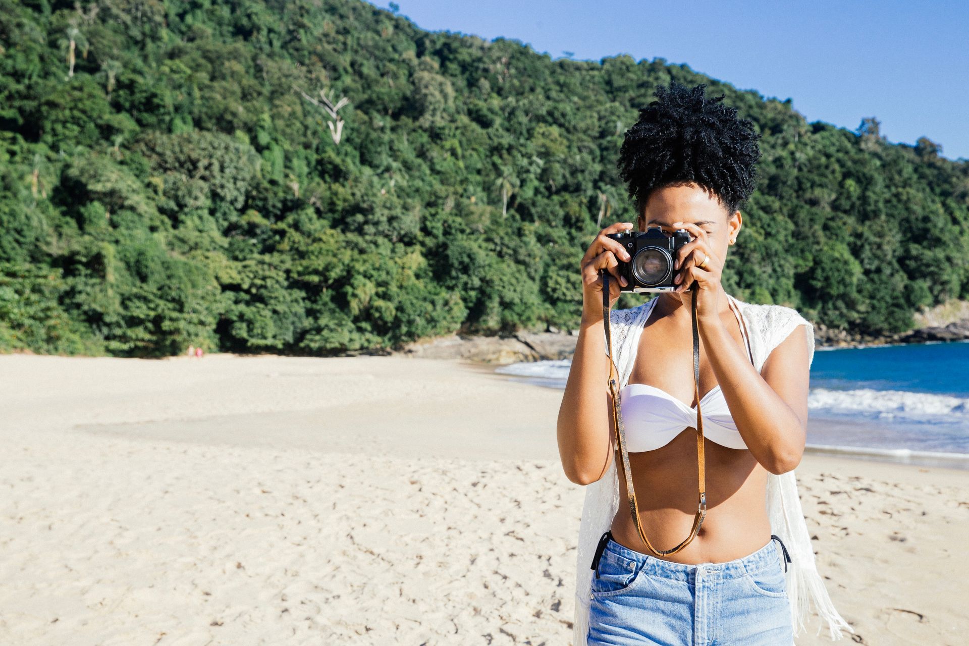 uma mulher negra com o cabelo black power amarrado segurando uma câmera tirando uma foto uma praia