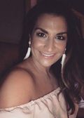 Woman with dark hair, wearing a blush off-shoulder top and white tassel earrings, smiling.