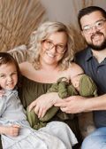 Family of four poses: parents, older child smiling, holding newborn in olive green, against a beige backdrop.