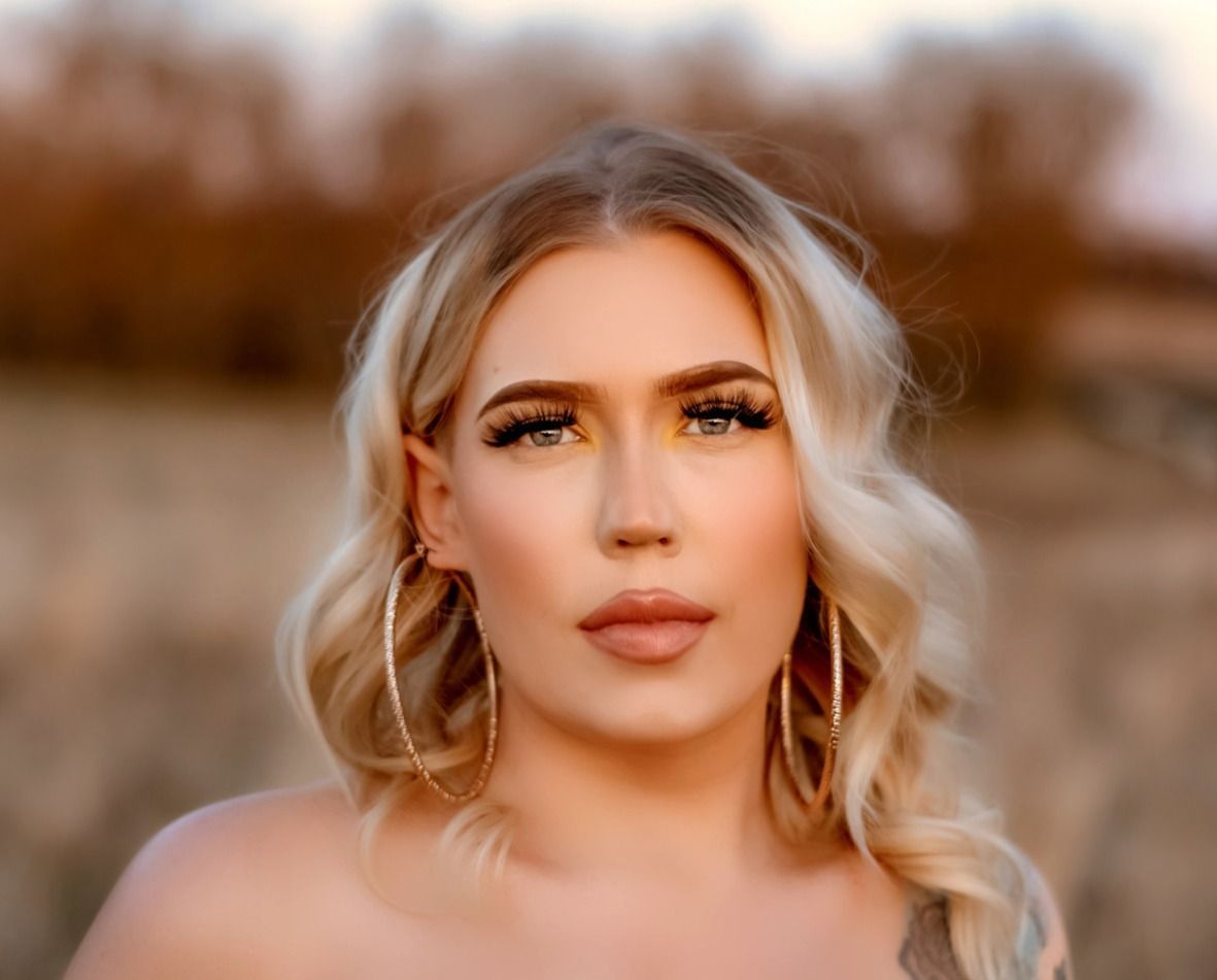 Woman with blonde hair and large hoop earrings poses outdoors, with a neutral expression.