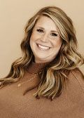 Woman with blonde highlights smiles, wearing a brown sweater against a neutral background.