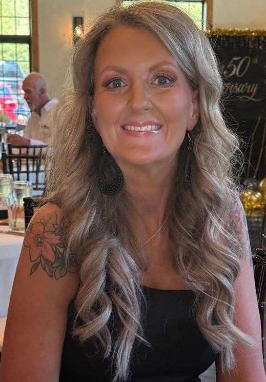 Woman with long, wavy blonde hair smiles, wearing dark top, sitting inside near decorated background.