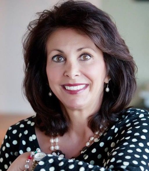 Woman in black polka dot top smiles, arms crossed, resting on chair.