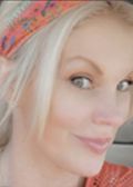Blonde woman with a patterned headband smiles; soft lighting, outdoors.