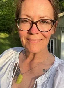Woman with dark hair, wearing a blush off-shoulder top and white tassel earrings, smiling.