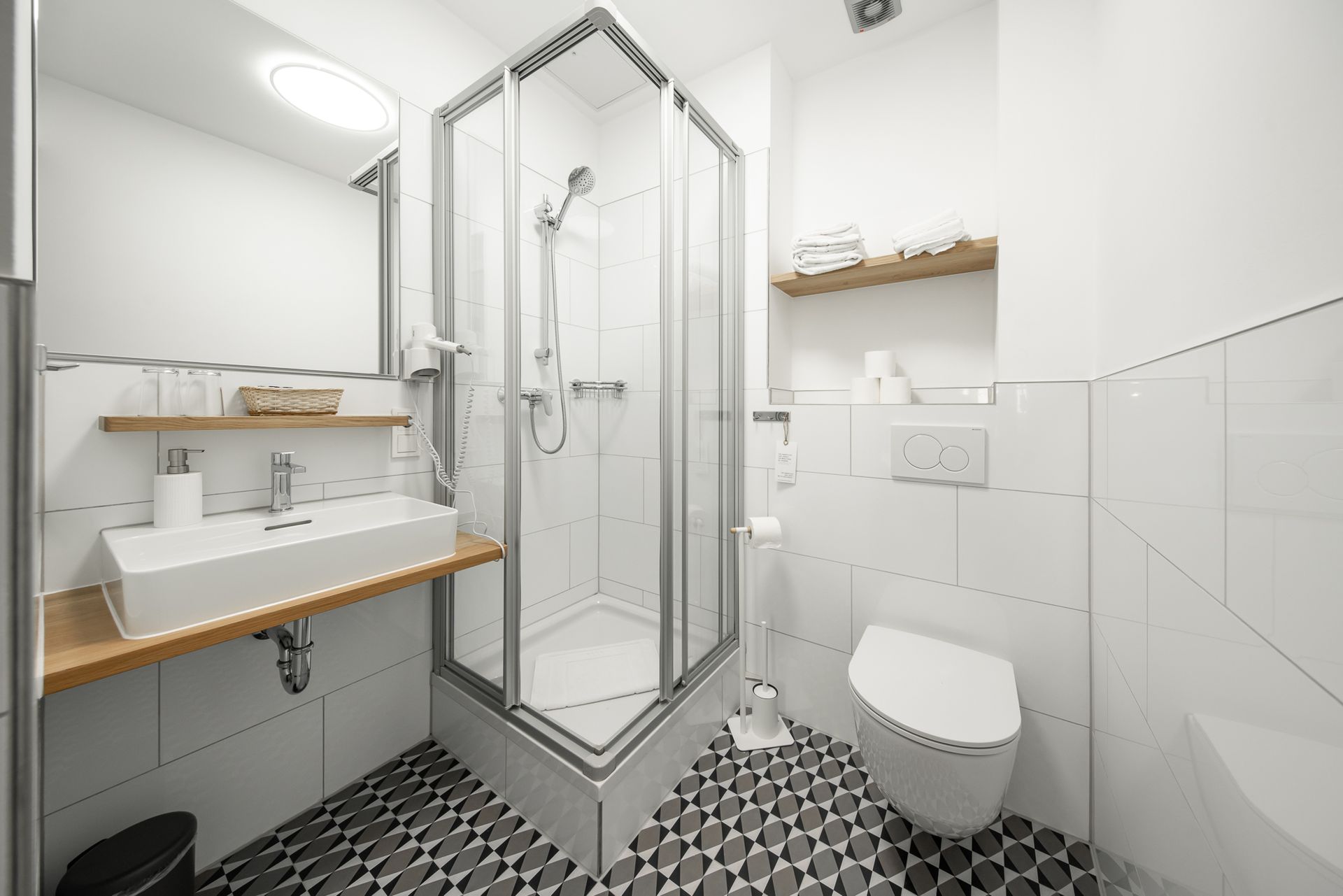 Small, white bathroom with a corner shower, floating sink, and toilet, with a patterned floor.