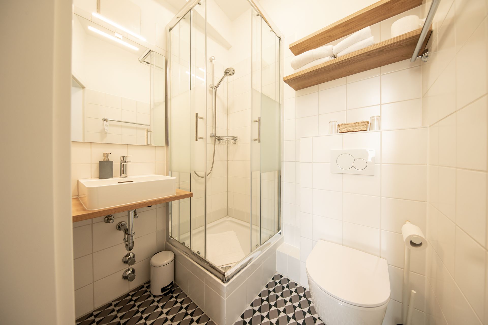 Small, white bathroom with a glass shower, toilet, sink, and two wooden shelves with towels.