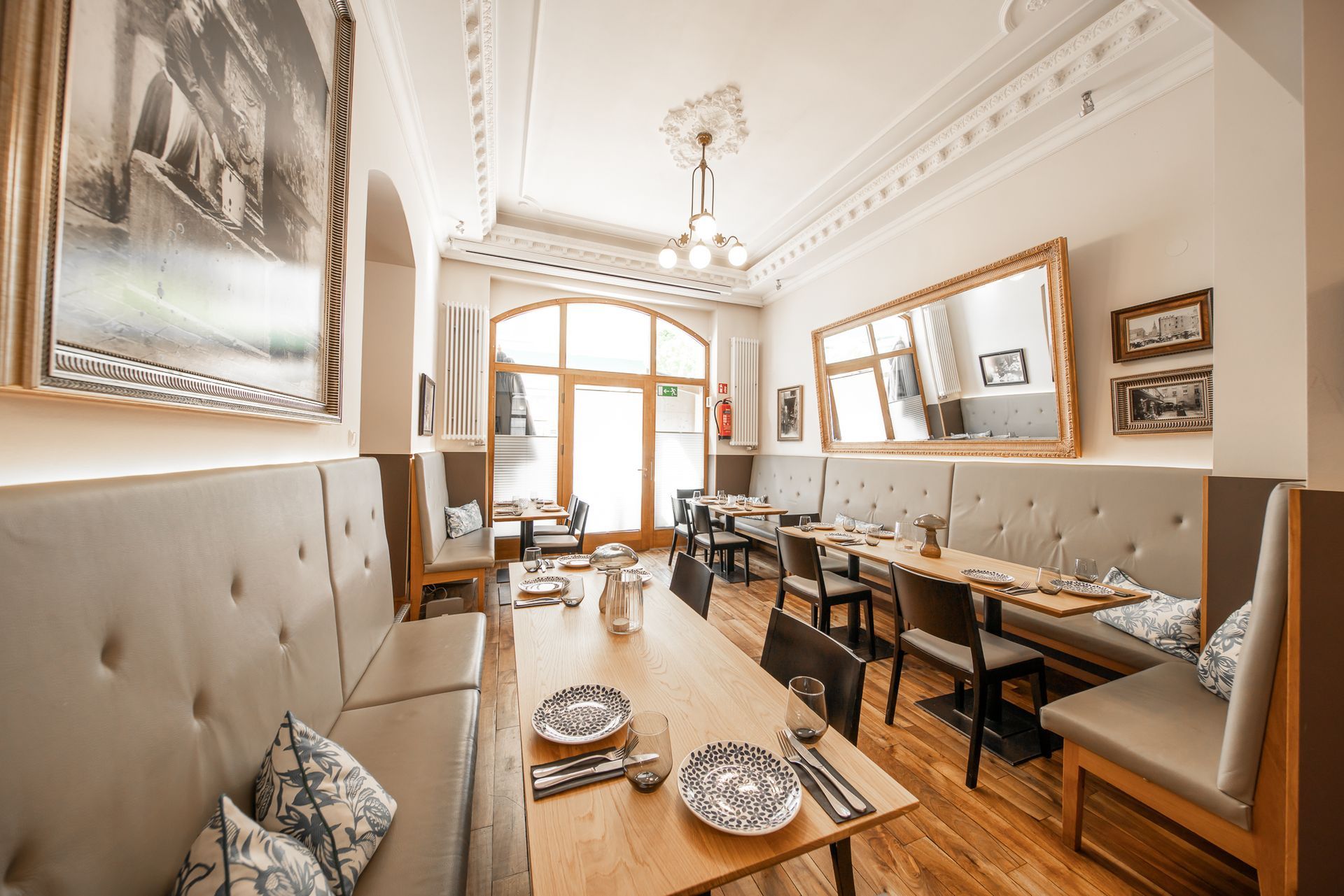 Restaurant interior with long wooden tables, booths, and large mirror. Bright room with decorative molding and artwork.