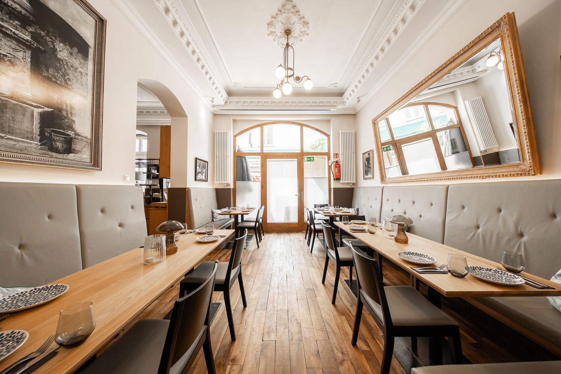Restaurant interior with long wooden tables, upholstered seating, large mirror, and open doorway.