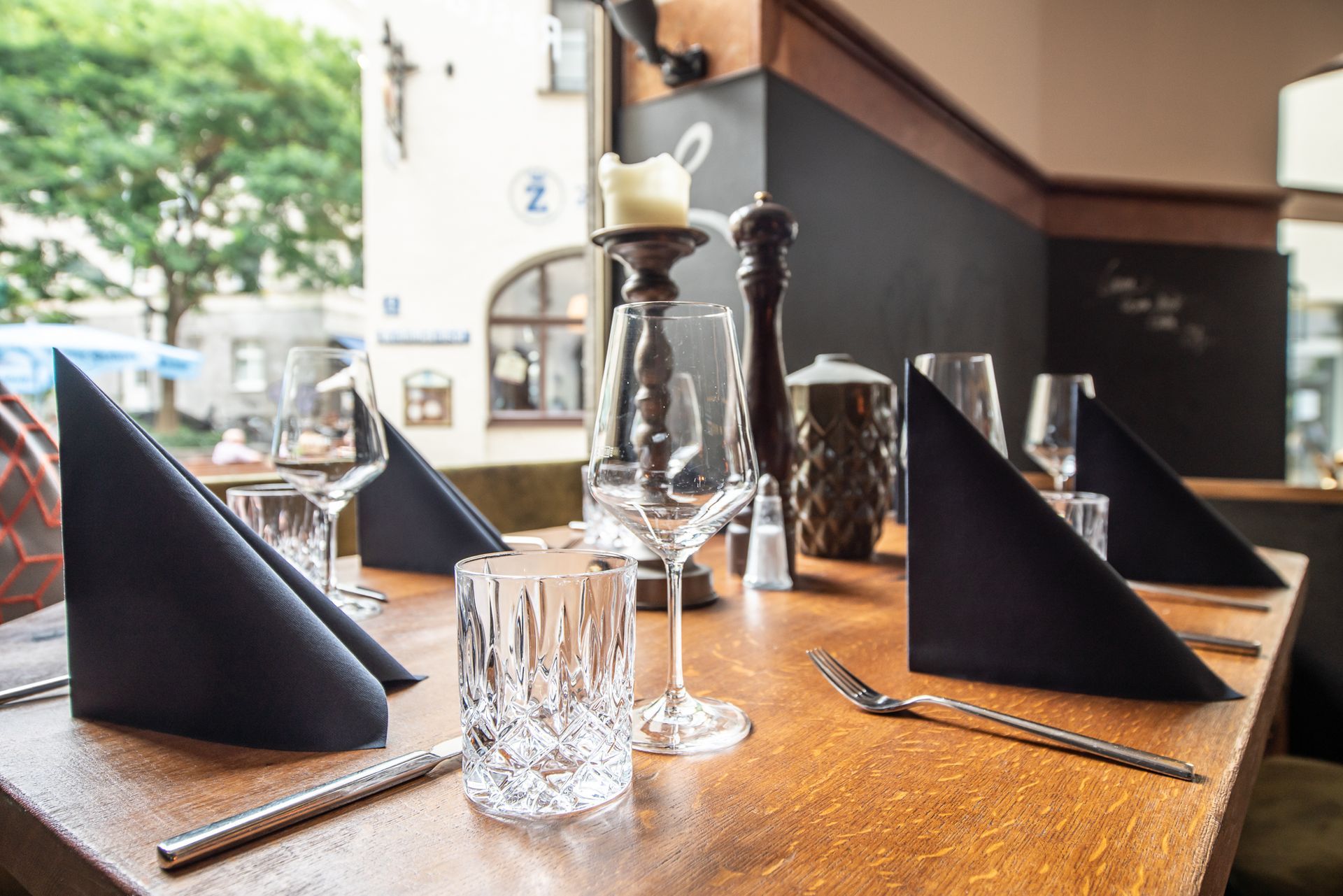 Restaurant table set with black napkins, glassware, and candle holder, near a window.