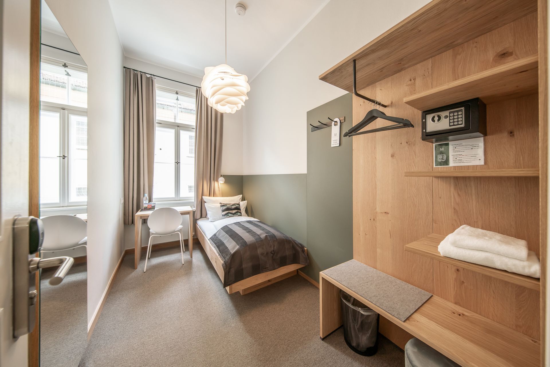 Hotel room with single bed, desk, wooden closet, and window. Green and white walls, neutral tones.
