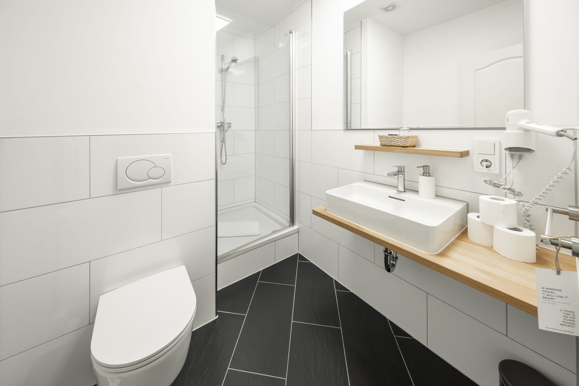 Modern white bathroom with black tiled floor; toilet, shower, sink, and mirror.