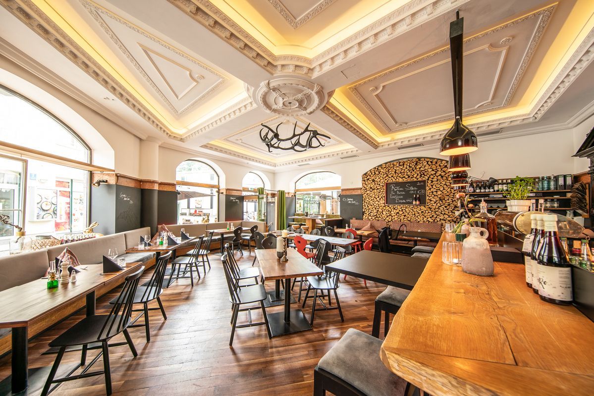 Interior of a restaurant with tables, chairs, bar, and decorative ceiling lights. Wooden accents and windows.