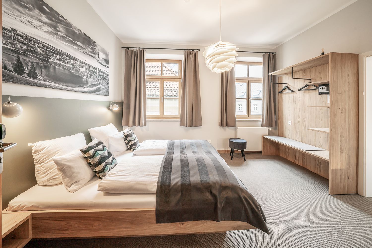 Hotel bedroom with a large bed, windows, and light wooden furniture.