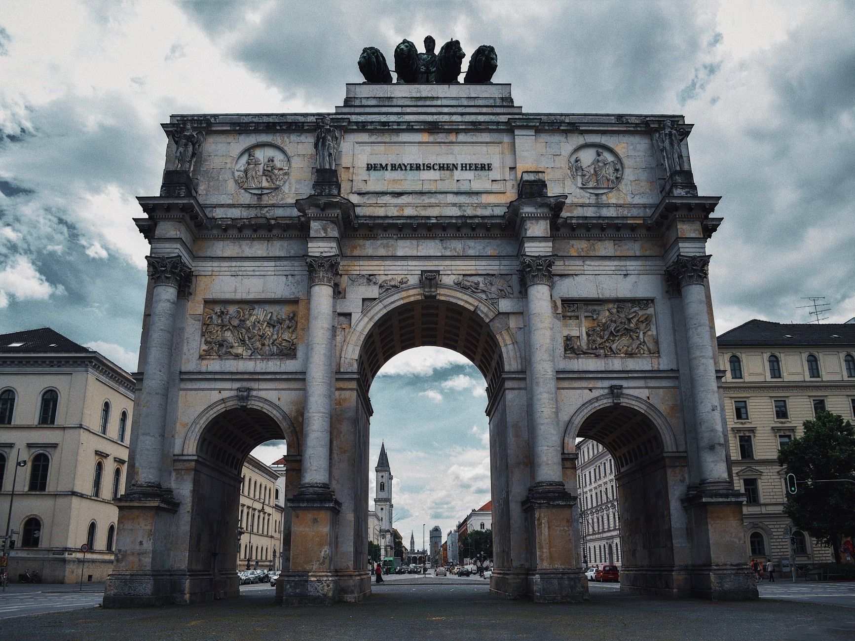 Triumphal arch with ornate carvings, overlooking a street and buildings; cloudy sky.