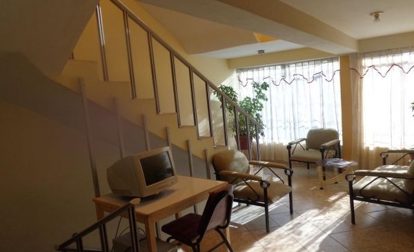 Sala de estar con escalera, escritorio y televisor, sillas de mimbre y ventanas luminosas con cortinas.