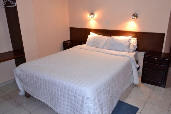 Cama doble bien hecha con edredón y almohadas blancas en una habitación con iluminación tenue.