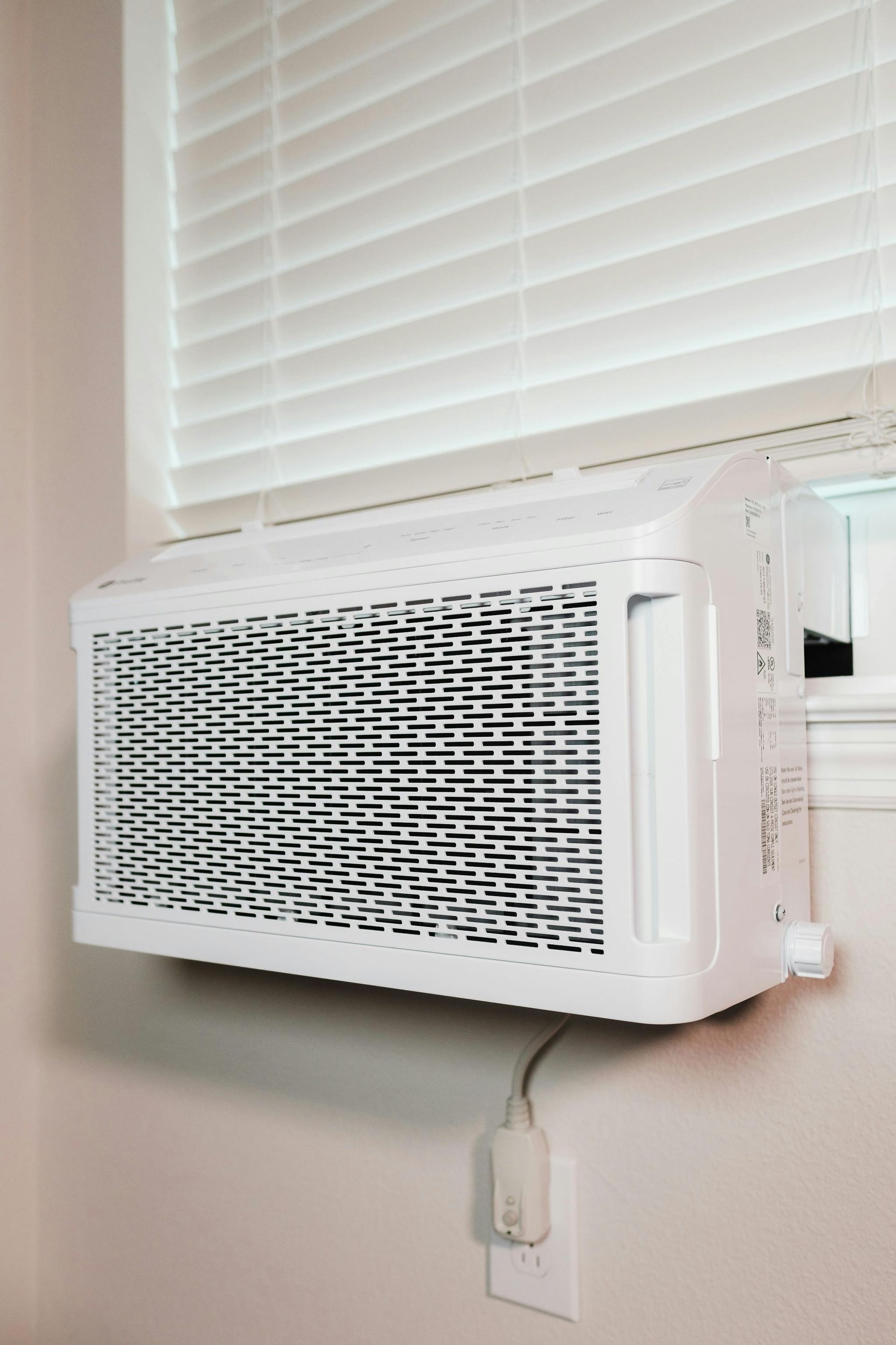White window air conditioner in a window with closed white blinds; power cord plugged into an outlet.