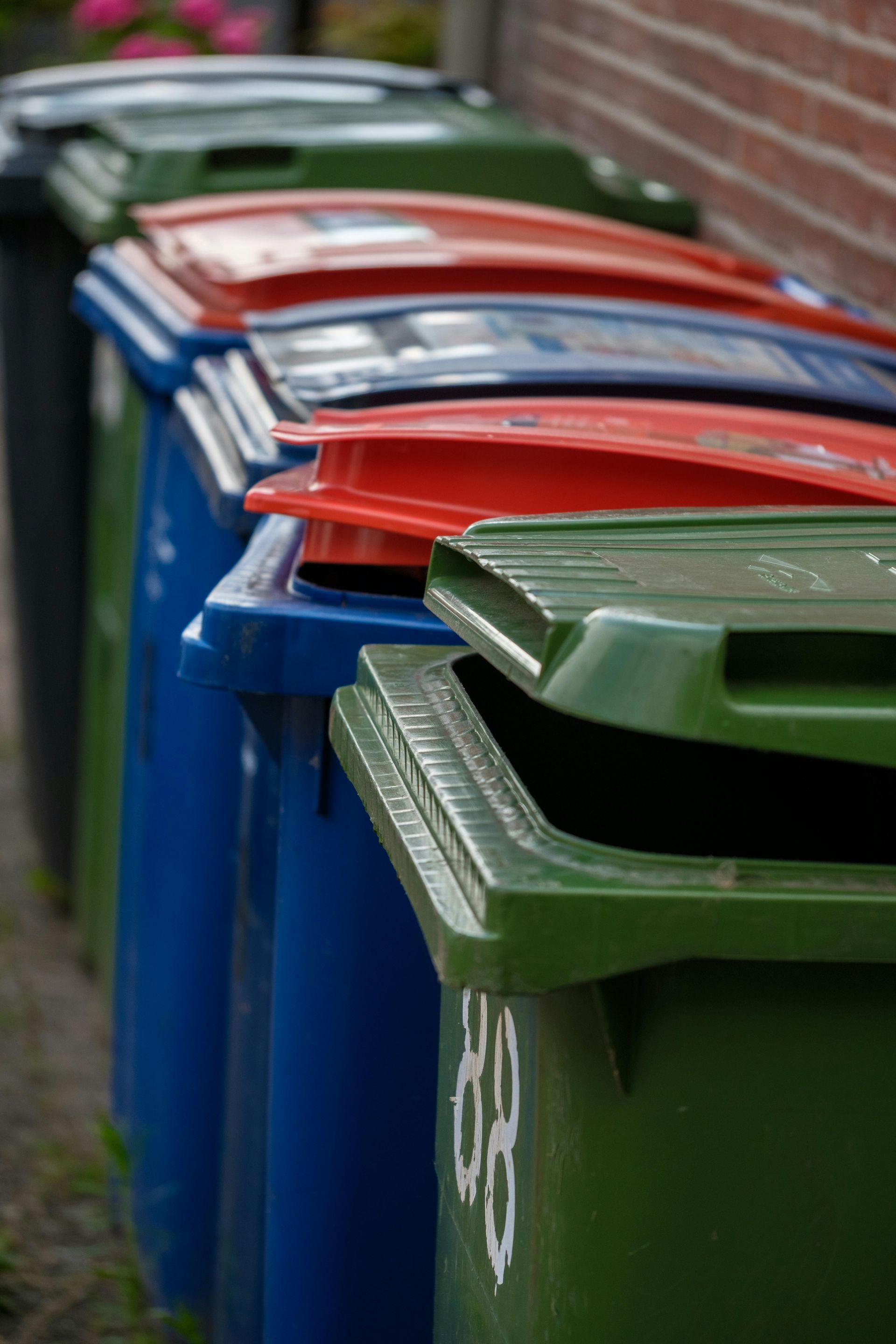Line of colorful recycling bins with red, blue, and green lids against a brick wall.